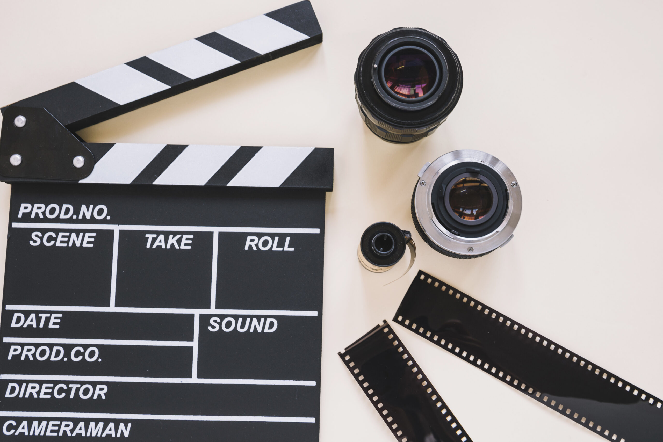 Foto de equipamentos audiovisuais, são eles: uma claquete, duas lentes e 3 filmes fotográficos em cima de uma superfície branca.