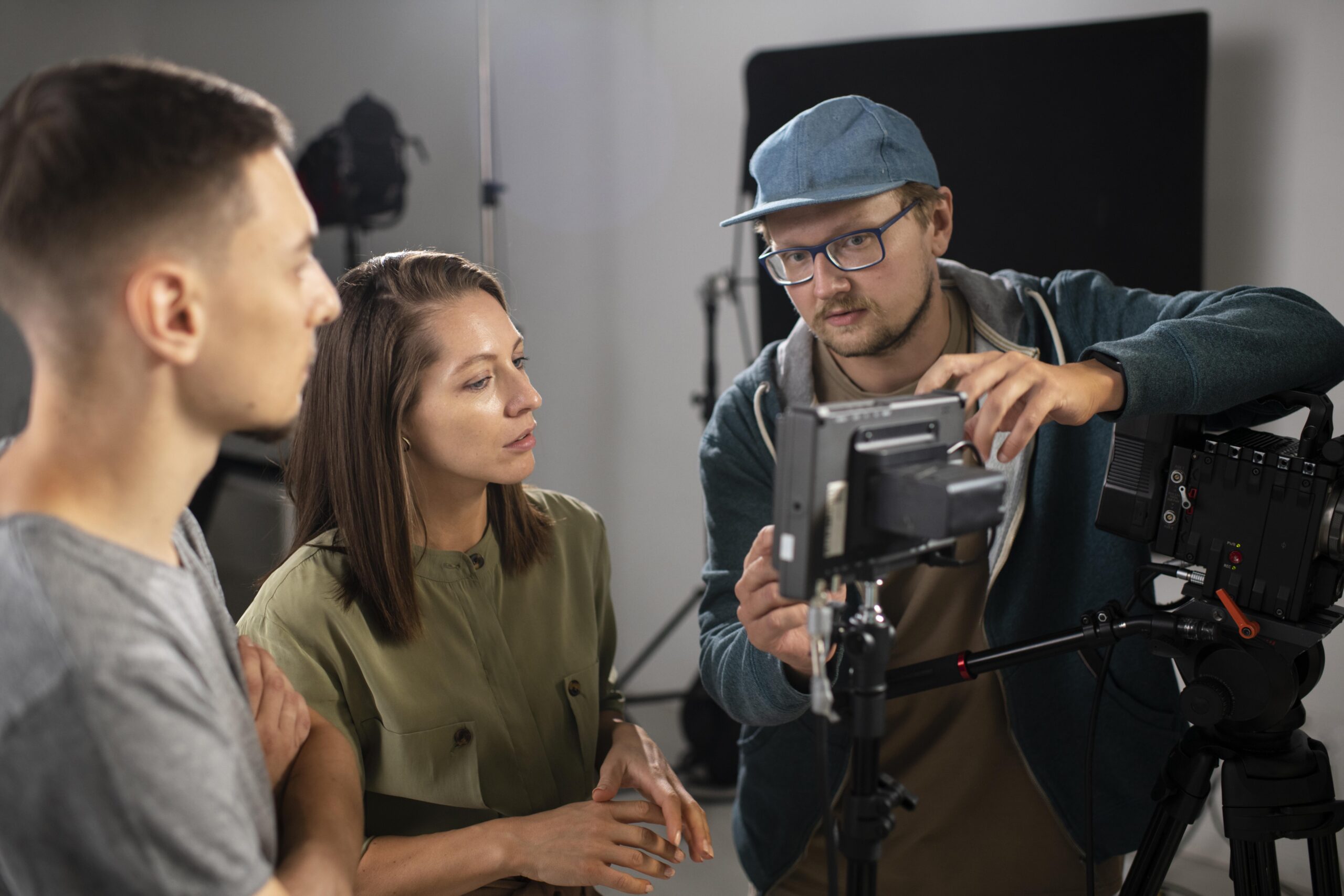 Foto de um estúdio audiovisual, no qual um produtor mostra para sua equipe de duas pessoas, uma mulher jovem de cabelos no ombro e blusa verde, e um homem jovem de camiseta cinza, qual foi o resultado de uma captação em uma câmera.