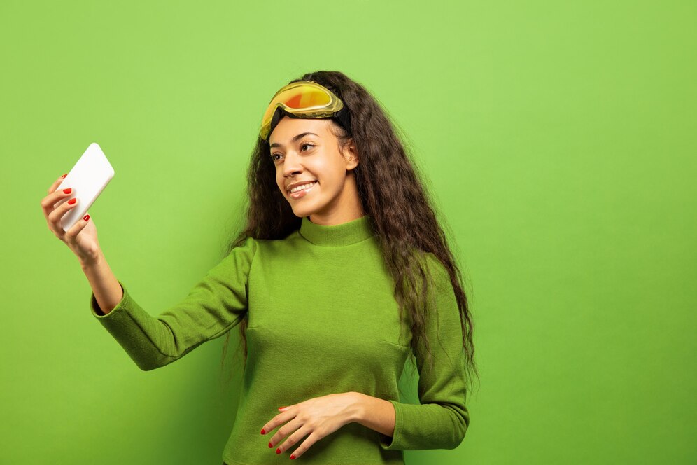 Mulher sorridente tirando uma selfie em frente a um fundo verde, usando blusa verde e óculos de proteção amarelo, ideal para aplicação de efeitos de chroma key.