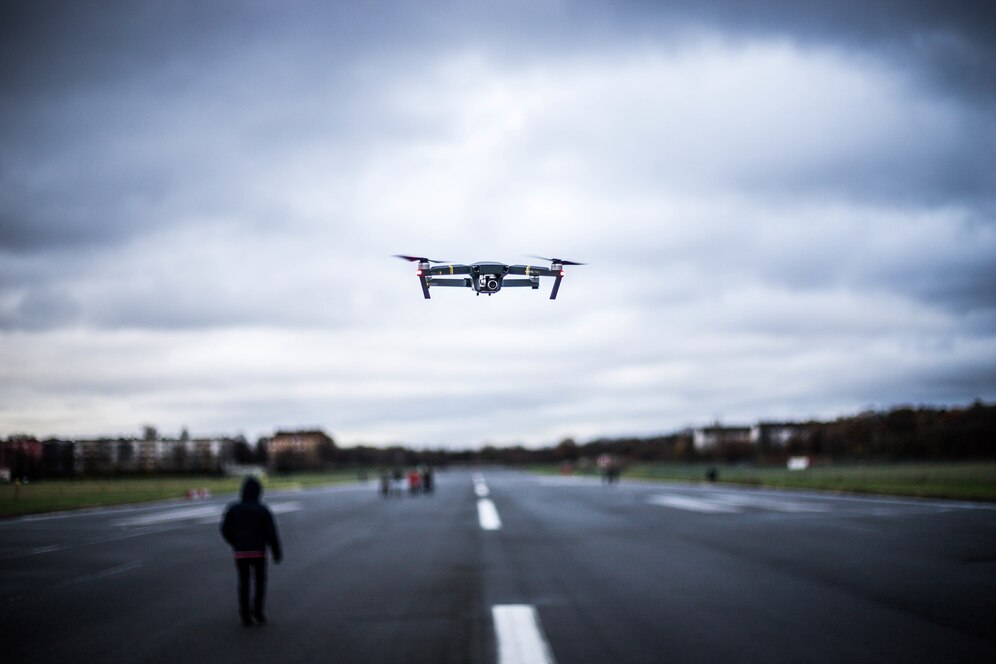 Drone capturando imagens em uma pista ao ar livre sob um céu nublado, enquanto pessoas caminham ao fundo, ilustrando o uso de tecnologia avançada na cobertura de eventos.