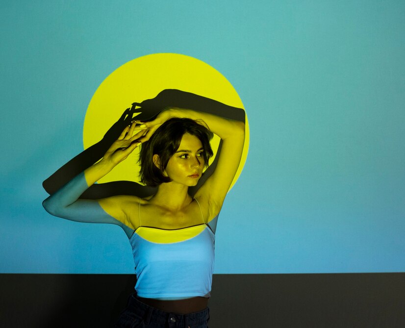 Fotografia conceitual de uma jovem de cabelo curto que está sentada com os braços levantados sobre a cabeça. Ela está posicionada contra um fundo azul-turquesa, com um círculo amarelo vibrante atrás dela.