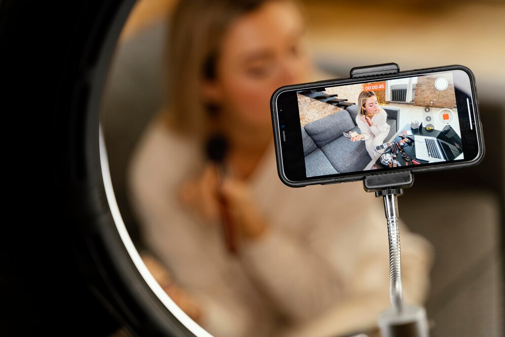 Close-up de um smartphone em um tripé, mostrando uma mulher aplicando maquiagem em um vídeo. A mulher que está gravando está levemente desfocada em segundo plano.